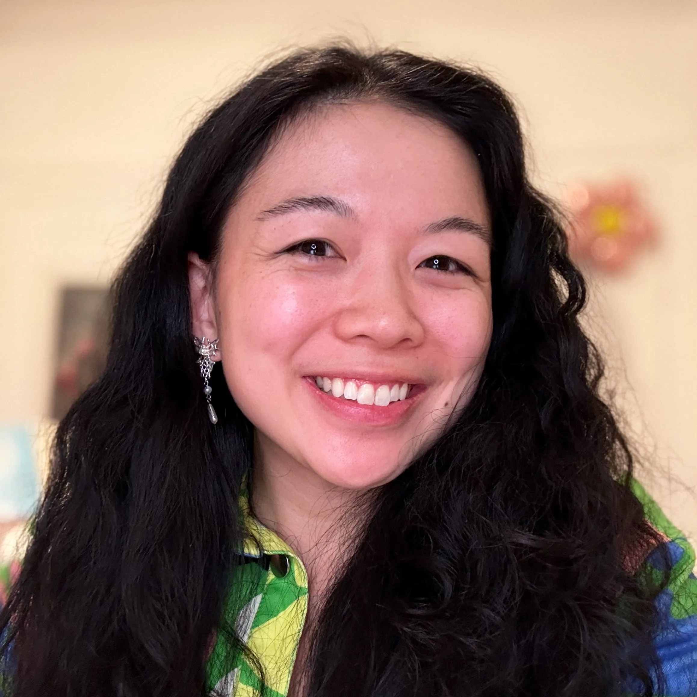 Smiling woman with long curly black hair wearing a colorful shirt and a dangling earring, with a blurred background.