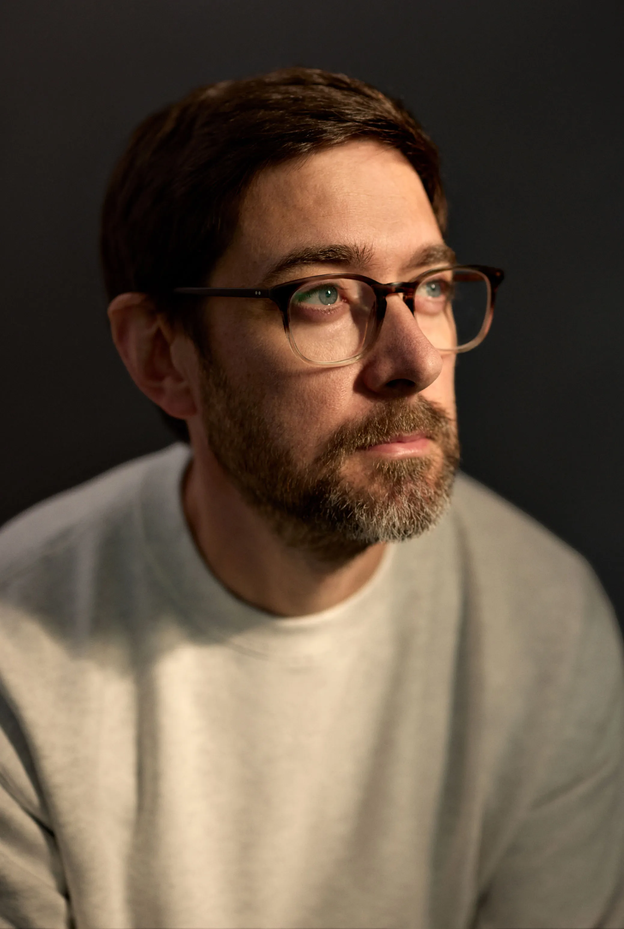 Portrait of a man with short brown hair, a beard, and glasses wearing a light gray sweater looking slightly upward with a thoughtful expression against a dark background.