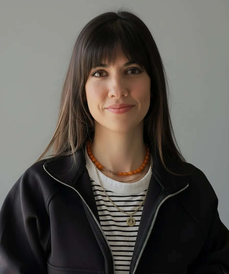 Portrait of a woman with long dark hair and bangs, wearing a black jacket over a white and black striped shirt, and two necklaces, one with orange beads and one with a gold pendant, smiling gently against a plain gray background.