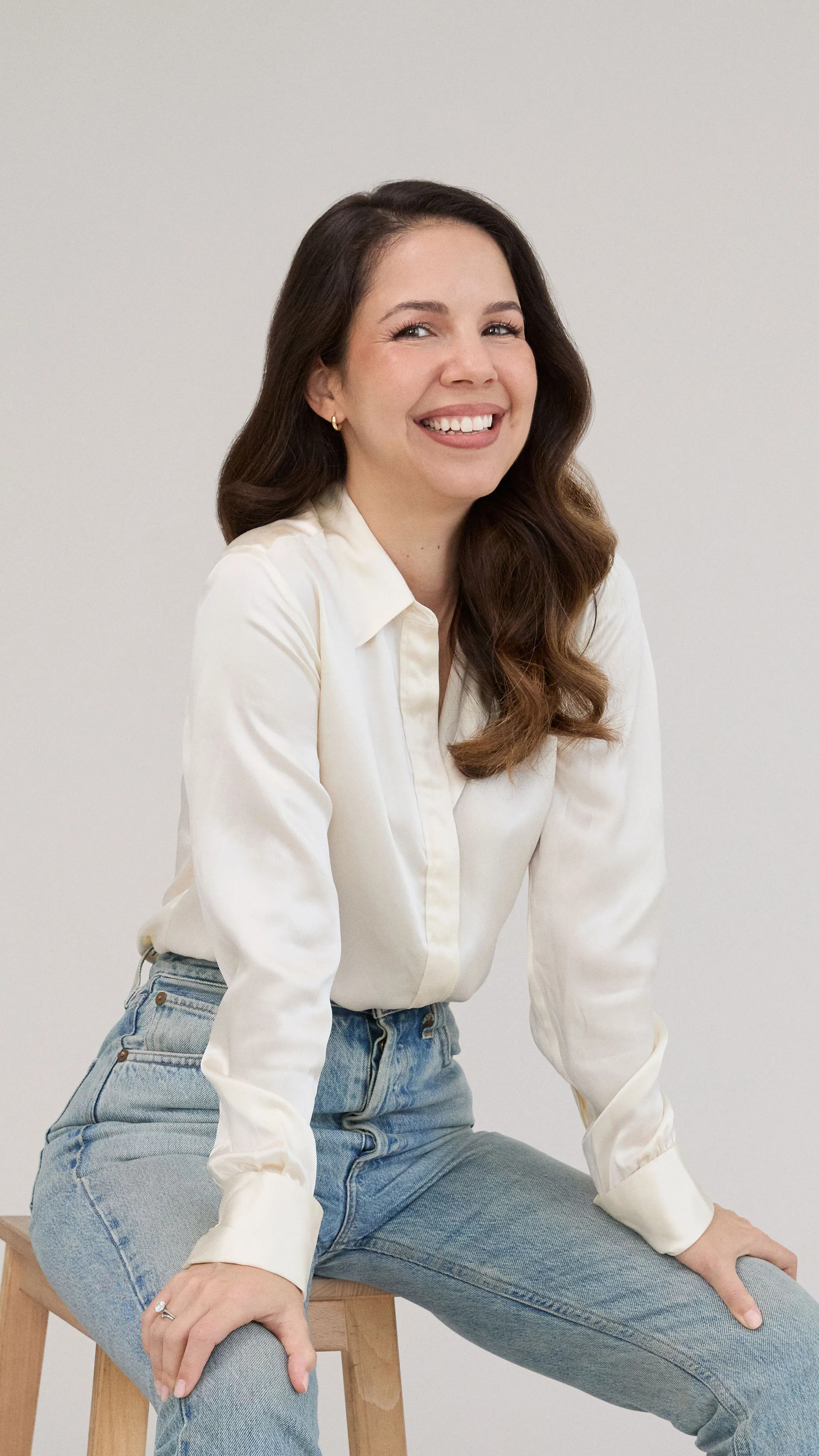 Smiling woman with long wavy brown hair wearing a white blouse and light blue jeans, sitting on a wooden stool against a plain light background.