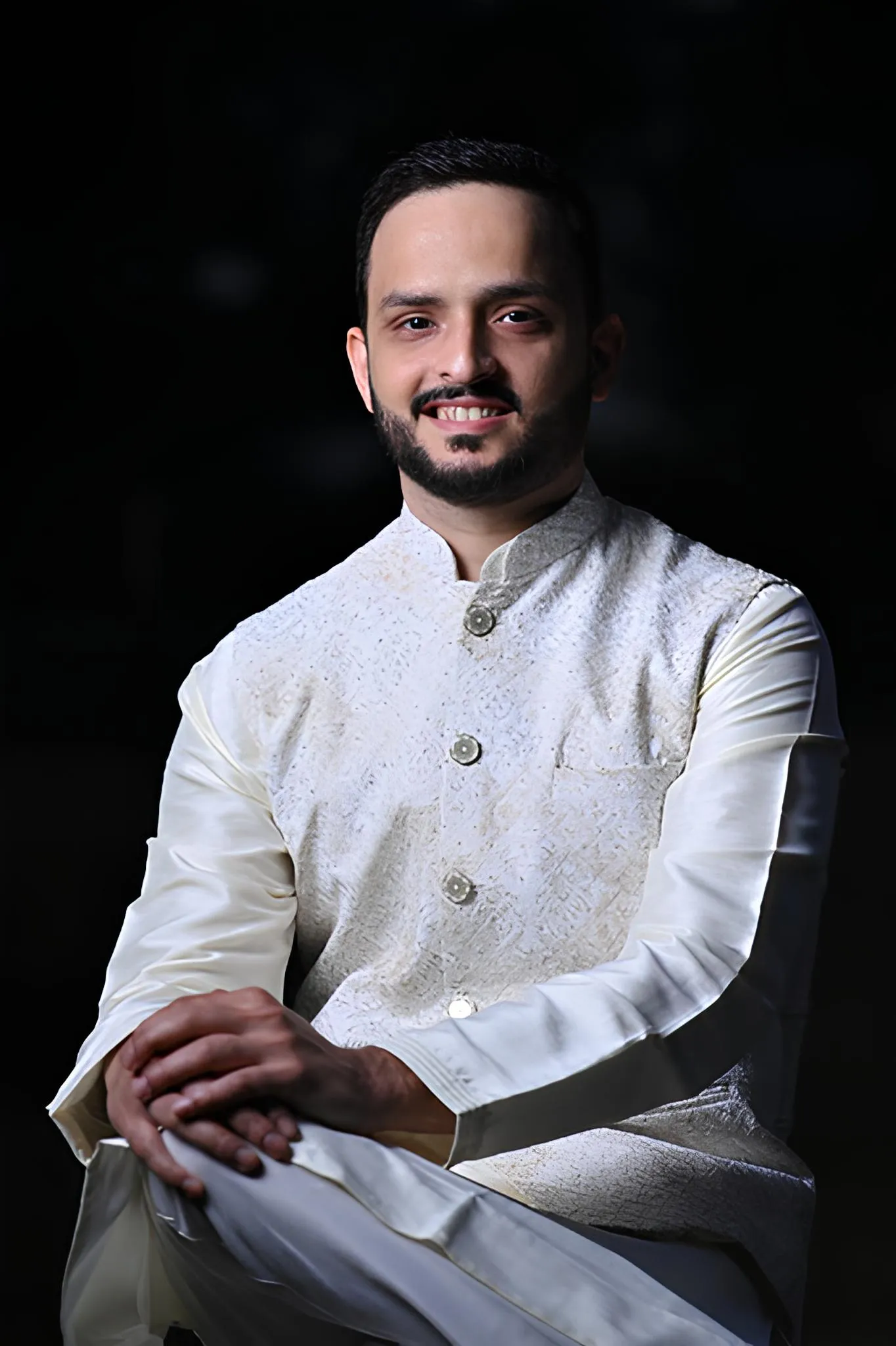Man with short black hair and beard wearing a traditional white embroidered kurta and smiling against a dark background, seated with hands clasped on his knee.